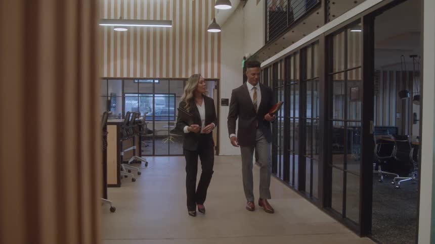 Two colleagues a young man and a young woman dressed in business attire walking down an office hall toward a conference room