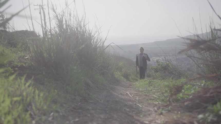 A man using trekking poles hikes up a dirt path on a grassy hillside walking towards the camera on a bright and hazy day