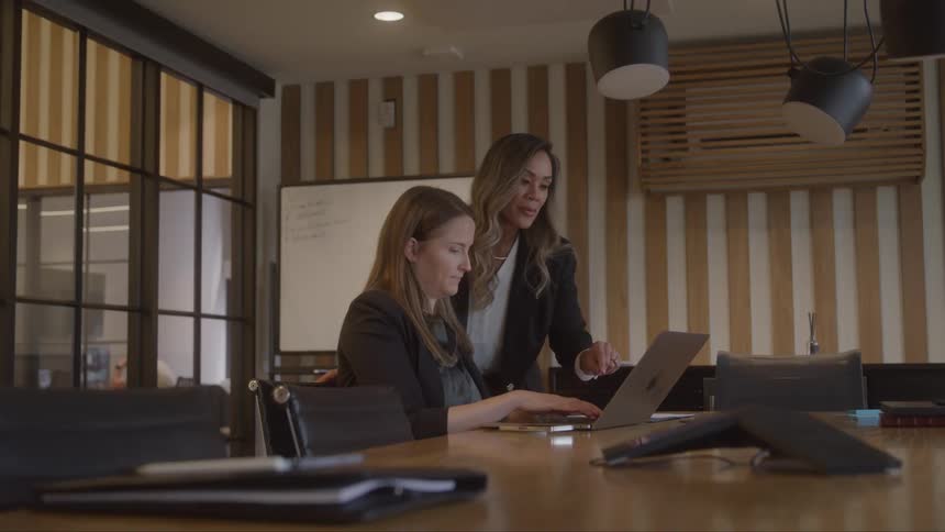 Two women are collaborating at a conference table in an office one woman who is seated works on a laptop while the other stands beside her pointing at the screen and discussing something