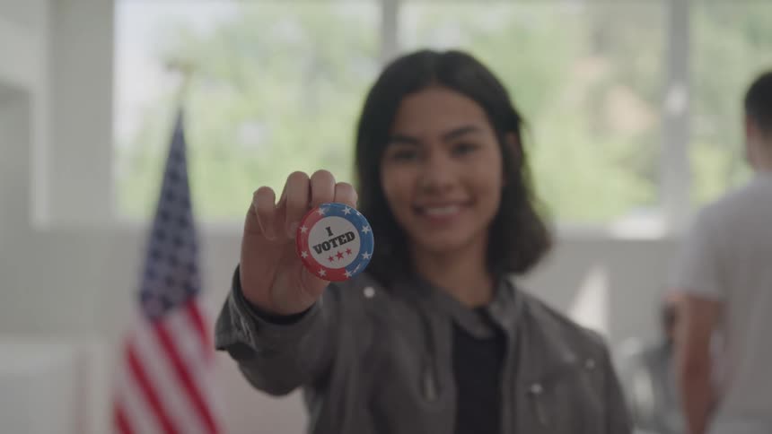 A young woman in front of an american flag proudly shows off her button with the words i voted