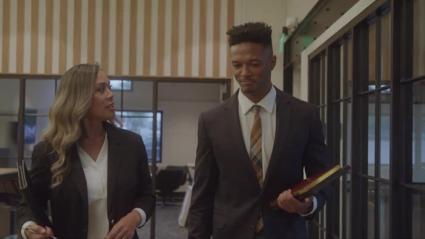 A male and a female colleague are walking and talking in an office hallway the man is holding a folder and looking at the woman as she speaks to him they are both dressed in professional business attire