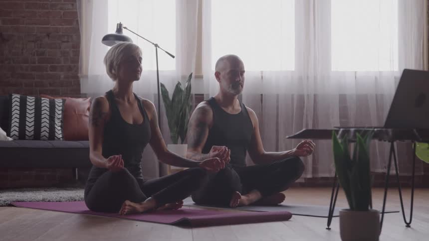 Two people sitting in meditation pose on yoga mats in front of a laptop