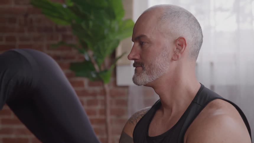 A man with a shaved head and a gray beard looks to the right in a profile view as he sits next to a woman wearing black clothing who is also working out
