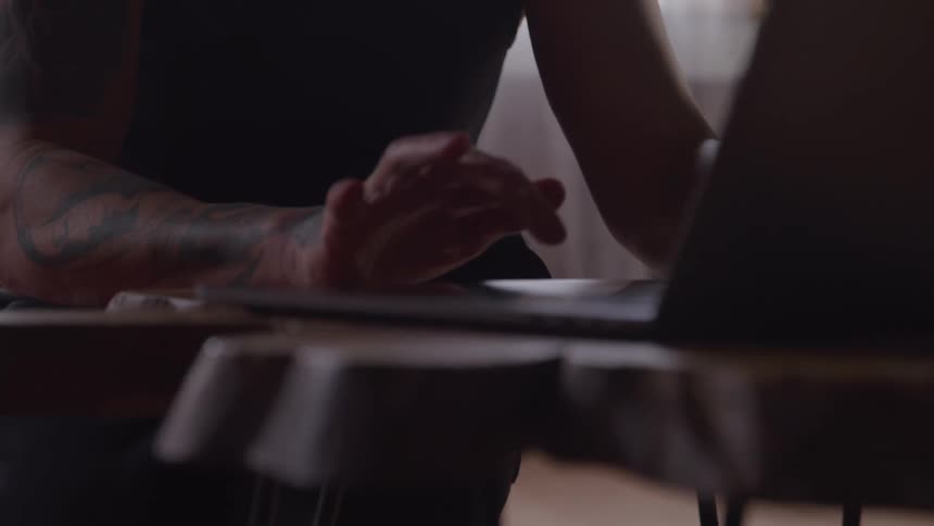 A person with tattoos on their arm is working at a desk looking at a laptop computer they are shown from the chest up and the focus is on their hands as they type and use the touchpad