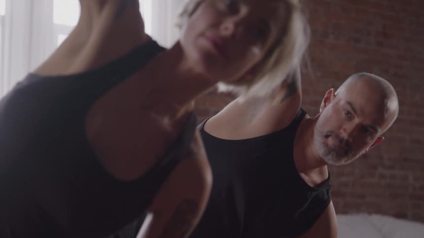 A man and a woman stand together in a room with a brick wall both wearing black workout clothes and doing a yoga pose with one arm raised in the air