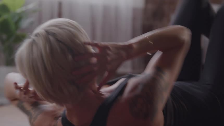 A blonde woman is exercising on a purple yoga mat doing crunches with her hands behind her head she is wearing a black sports top and has tattoos on both arms