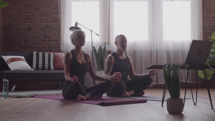 Two people in meditation pose on yoga mats in front of a laptop