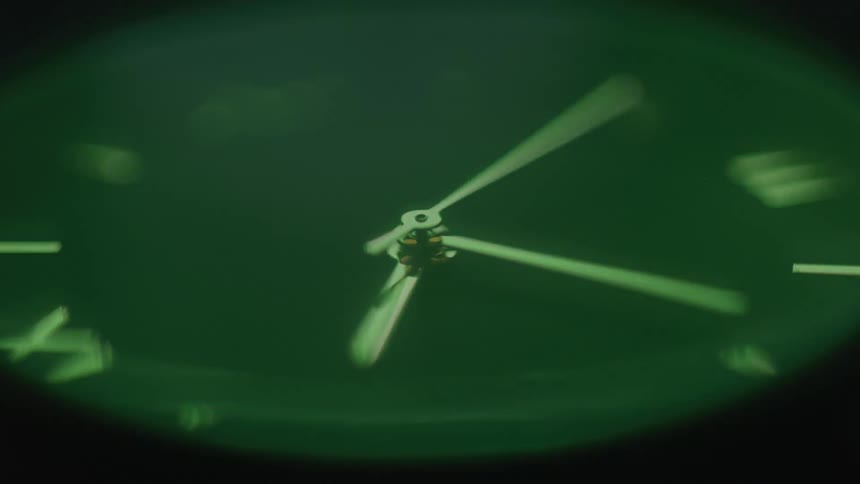 A green watch face with white hands and markers is blurred with the second hand rapidly sweeping around the dial