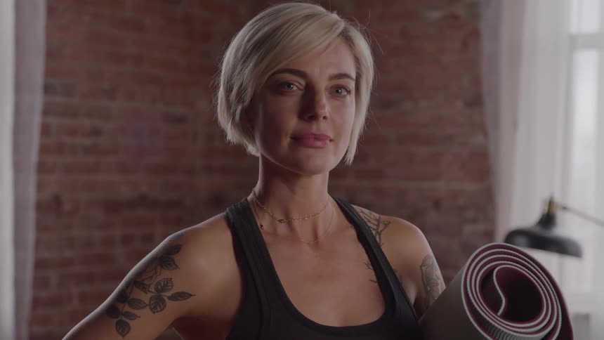 A woman with short blonde hair and tattoos on her arms holds a rolled up yoga mat under her left arm looking directly at the camera with a neutral facial expression
