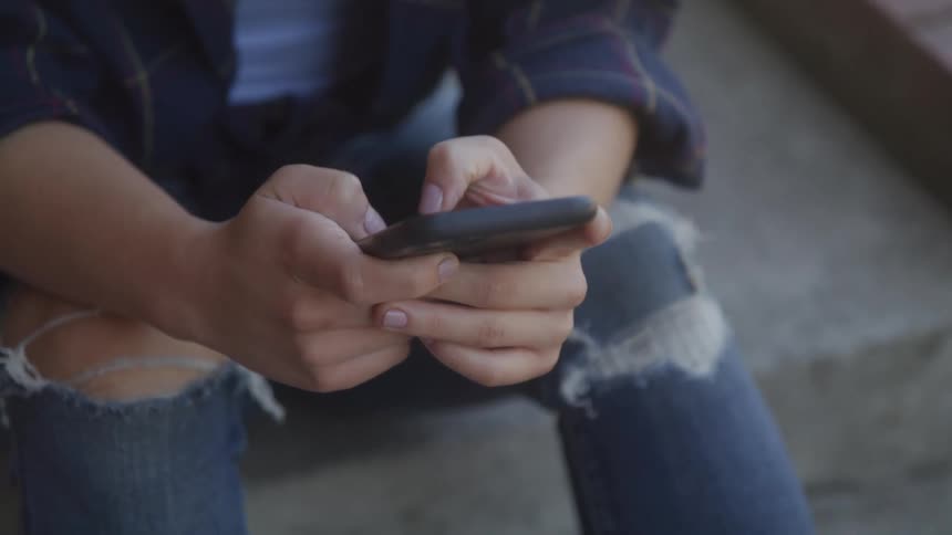 A closeup shot shows a persons hands holding a smartphone while their fingers type the person is wearing ripped jeans and a plaid shirt