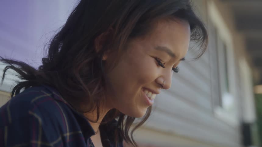 A person with dark hair wearing a plaid shirt and a silver necklace is looking down with a neutral expression their face then breaks into a wide smile as they laugh