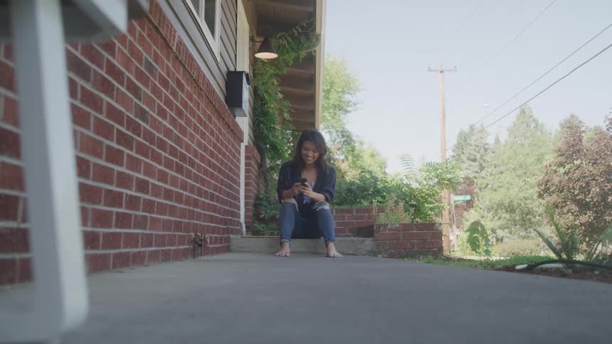 A barefoot woman relaxes on her front stoop smiling at her phone