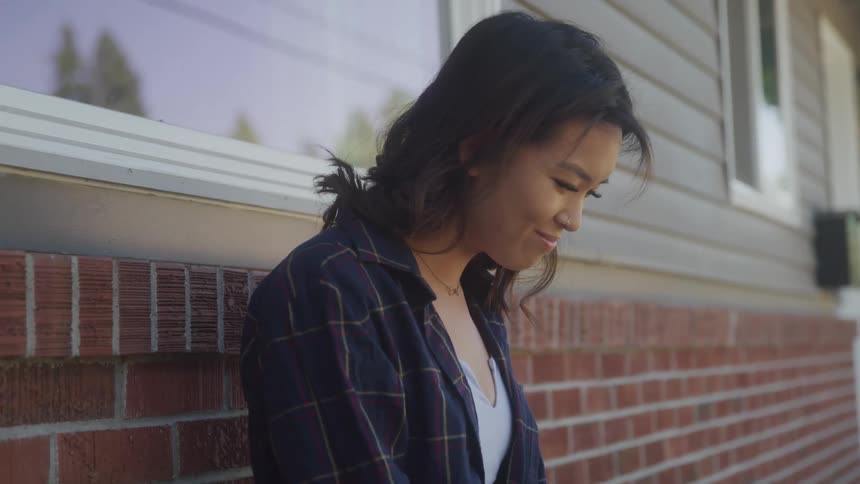 A young woman is leaning against a brick wall outside a building with a white siding she is smiling gently and looking down as a warm glow from the sun highlights her hair and face