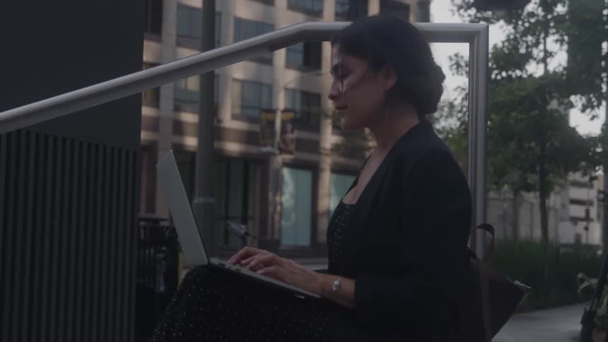 The woman types on her laptop while sitting on the steps outside the building