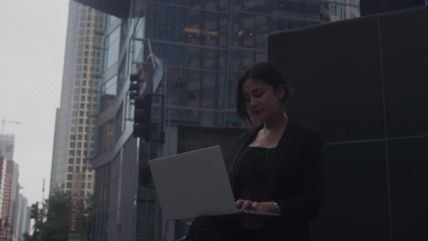 Woman using a laptop sitting in an urban area with mirrored buildings in the background