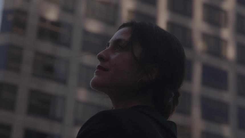 A woman with her hair pulled back and a dark shirt stands outdoors looking up toward the top of a modern building with a pattern of square windows