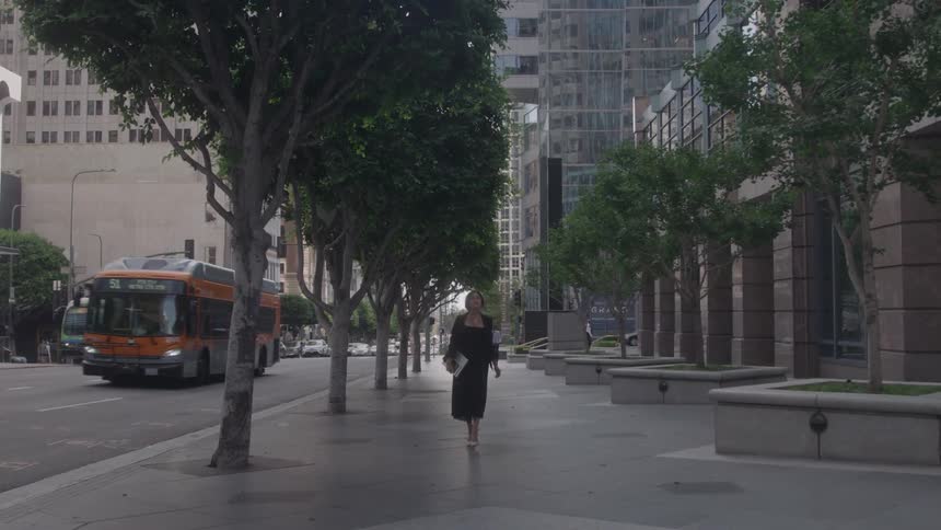 A woman in a long black dress and heels walks toward the camera down a city sidewalk lined with trees and modern buildings as a bus and cars drive on the street behind her