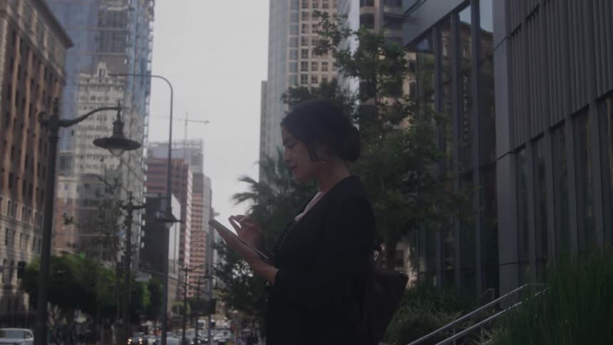 A young woman standing on the sidewalk scrolling through her phone in the city at midday