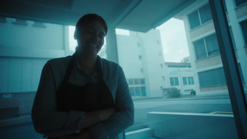 A chef in an apron smiles while standing near a window