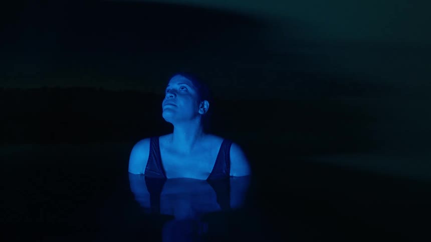 A woman stands still in a lake at night looking around in fear