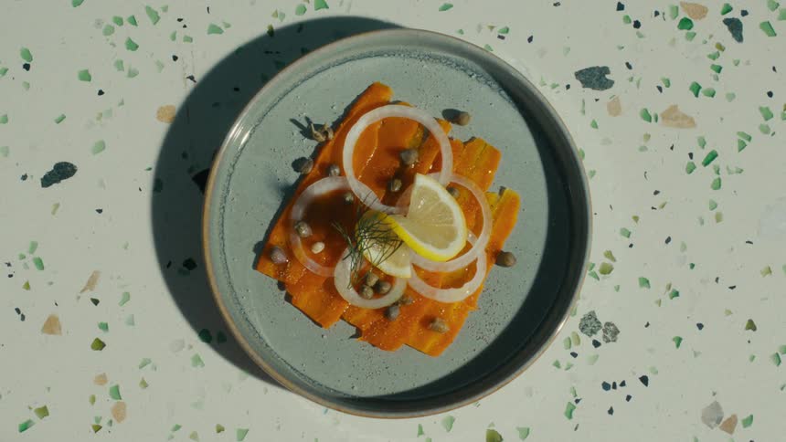 A hand serves a plated dish of carrot lox with garnishes on a grey plate