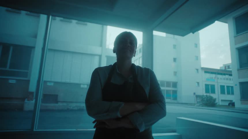 Person wearing an apron stands with arms crossed in front of a large window with buildings in the bluish background