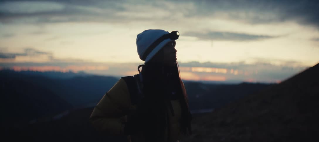 A woman carrying a backpack and wearing a cap and a lantern on her head stands on the slope on the side of a mountain looking at the way up