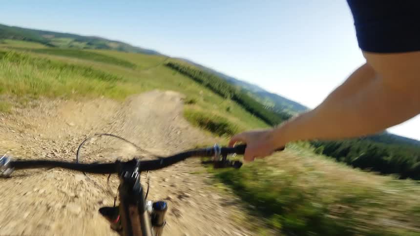 A cyclist rides down a bumpy dirt trail with the handlebars and hands of the rider visible in the foreground and a wide green and hilly landscape in the background