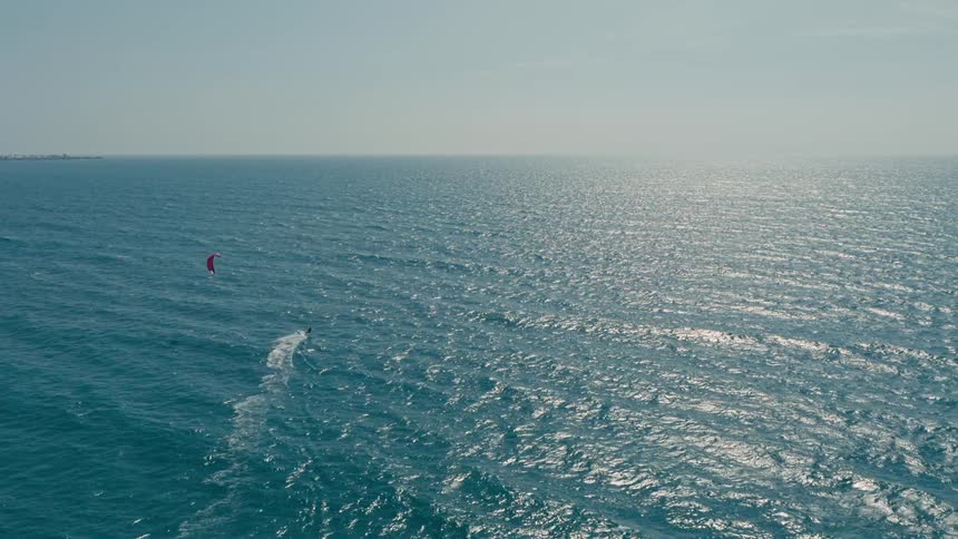 A wide aerial shot shows a person on a kiteboard gliding across the vast blue ocean the person with a red kite above them creates a foamy white trail behind their board