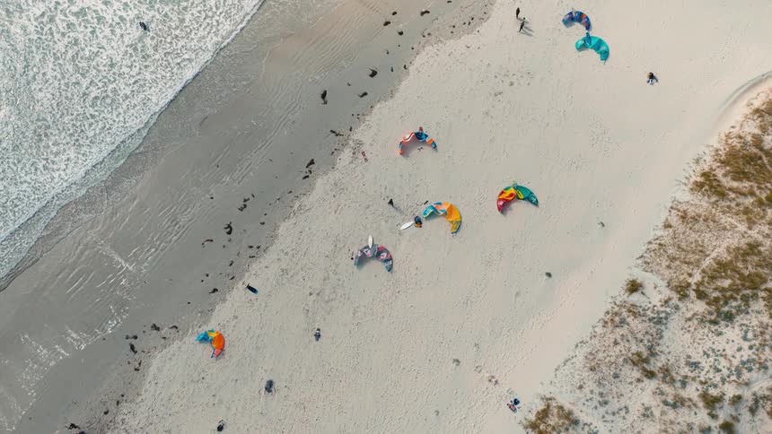 An aerial drone shot shows a sandy beach with several people kitesurfing