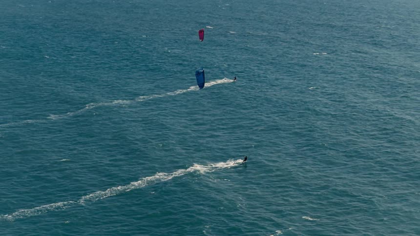 Three kite surfers each with a large colorful kite flying behind them travel across a wide blue body of water leaving thin white wakes behind them