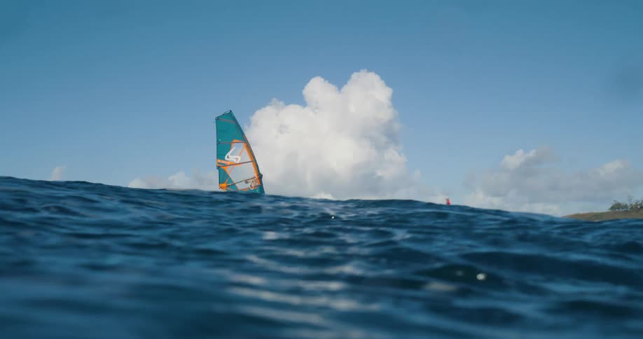 A windsurfer rides a large wave on a rough blue ocean with a large white cloud on the horizon and a distant shoreline visible on the right side of the frame