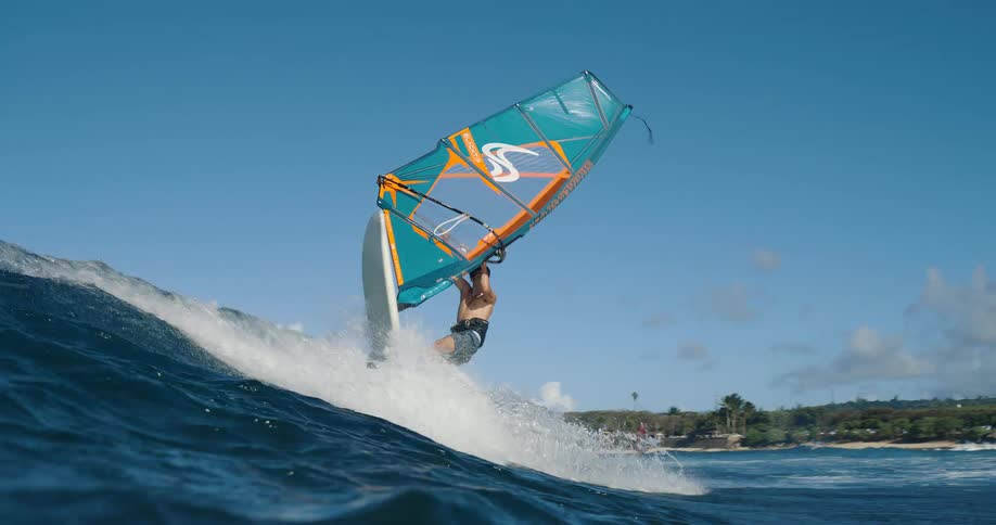 A windsurfer navigates a wave under a bright blue sky with scattered clouds