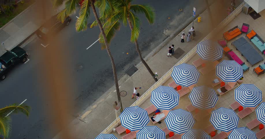 An overhead shot shows several blue and white striped umbrellas set up on a beach next to a road with palm trees people and cars can be seen moving around