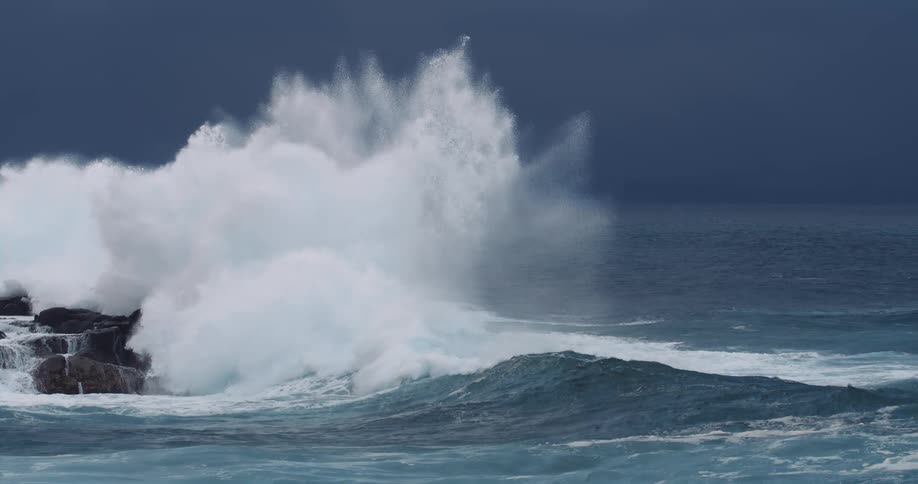Large waves crash against a rocky shoreline sending white spray into the air the water is a dark churning blue contrasting with the dark rocks and the stormy sky
