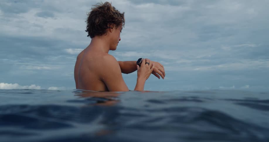 A man stands chestdeep in the calm ocean water under a cloudy sky looking down and adjusting the smartwatch on his wrist