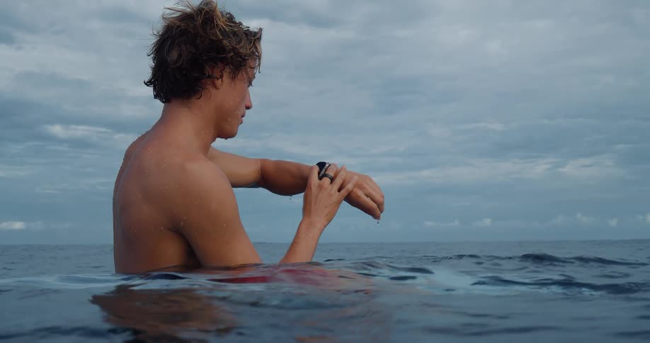 A young man is sitting on a surfboard submerged in the water and fiddling with his watch on his wrist
