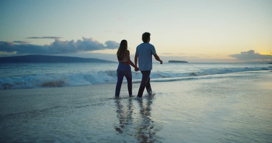 A man and woman holding hands as they walk on the edge of the water on a beach