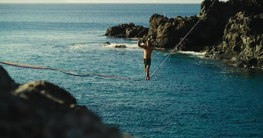 A man wearing only shorts walking on a high wire over the sea between two rock formations