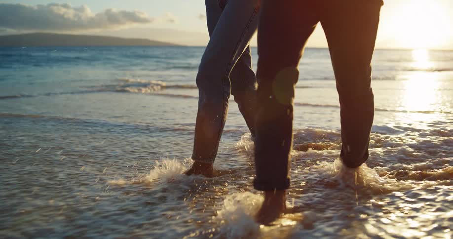 Two people walk on the beach as waves roll into shore and over their bare feet