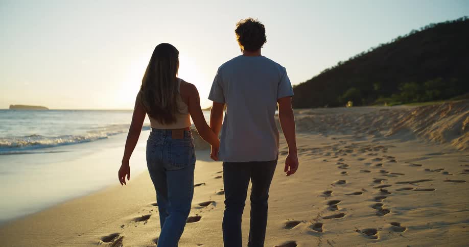 A man and woman smile and walk on the beach while holding hands