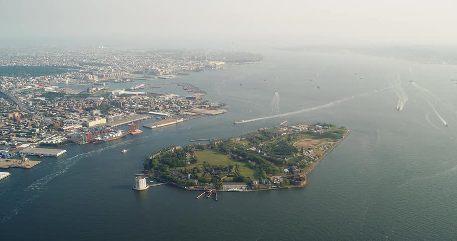 An aerial view captures a large green island in the middle of a bay the surrounding water is dotted with small boats and a sprawling coastal city is visible in the background