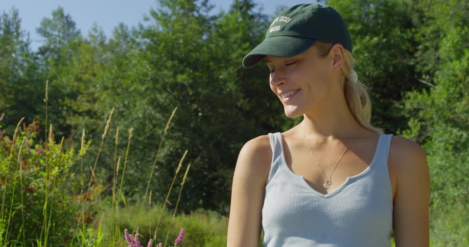 The video shows a blonde woman wearing a hat and a lightcolored tank top she is outdoors looking out at the scenery and then turns her head down and smiles