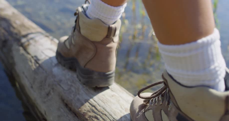 Person wearing hiking boots balances on a wooden pole lying in shallow water on a sunny day
