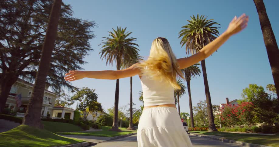 Happy blonde woman twirling while walking on residential street lined with palm trees
