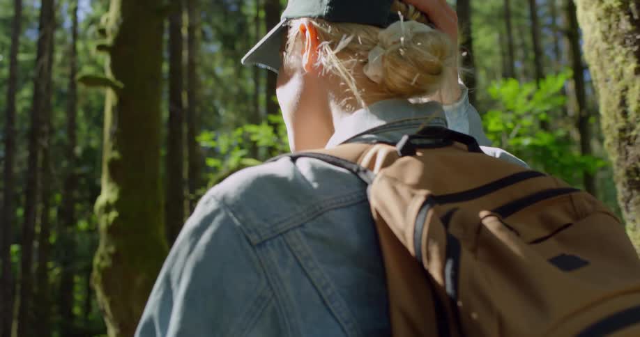 A woman wearing a backpack and a baseball cap is walking through a sun dappled forest