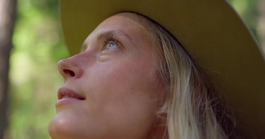 A closeup shot of a blonde woman wearing a hat looking up with a serene and joyful expression as she smiles