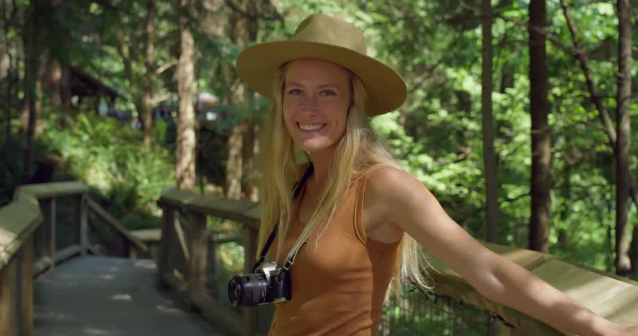 A woman with long blonde hair wearing a hat and a camera around her neck is walking on a wooden bridge through a forest and turns to smile warmly at the camera