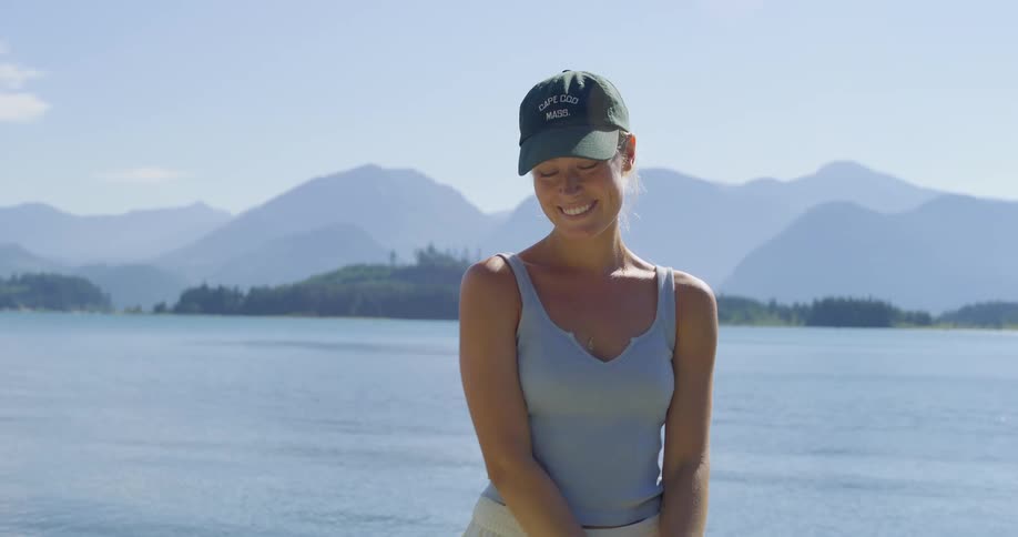 A woman wearing a baseball cap and a tank top stands by a lake with mountains in the background putting on her hat and smiling