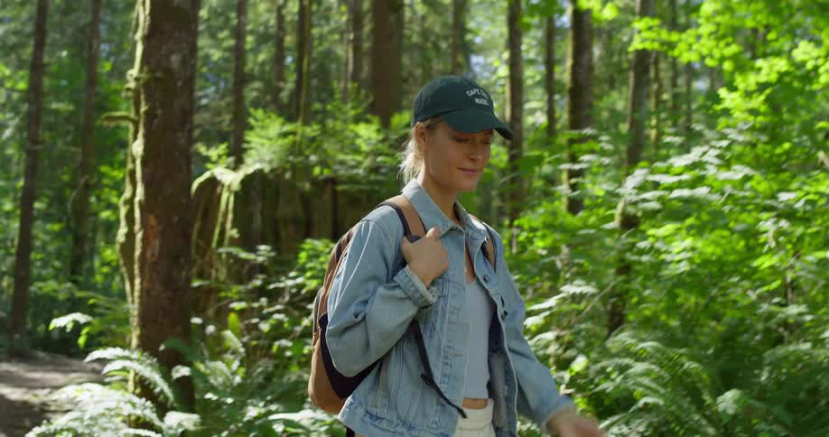Woman holds the straps of her backpack as she walks on a path through the forest looking at trees